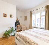 Világos hálószoba fa mintás padlóval, növényekkel és nagy ablakokkal egy kétszobás lakásban.