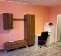 Garzonlakás bútorokkal, íróasztallal, mikrohullámú sütővel és fa mintázatú padlóval.