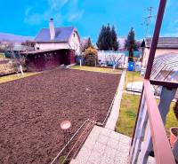 Rendezett kert a partizánskei ház mellett, magasított ágyásokkal és körülötte fákkal.