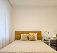 Hálószoba egy egyszobás lakásban világos ággyal, függönyökkel és fehér asztallal, lámpával.