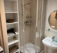 Fürdőszoba egy kétszobás lakásban sarok zuhanyzóval, mosdóval és fehér polccal.