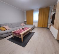 Nappali egy háromszobás lakásban kanapéval, függönyökkel és fa mintázatú padlóval.