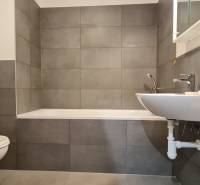 Fürdőszoba káddal, mosdóval és mosógéppel egy kétszobás lakásban. A szürke csempék uralják a teret.