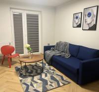 Nappali egy háromszobás lakásban kék kanapéval, piros fotellel és fa mintázatú padlóval.