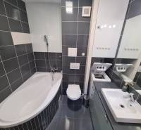 Fürdőszoba egy kétszobás lakásban káddal, mosógéppel, mosdóval és sötét csempékkel.