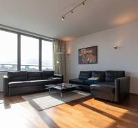 Nappali egy kétszobás lakásban fekete kanapéval, fa hatású padlóval és nagy ablakokkal.