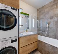 Fürdőszoba egy háromszobás lakásban mosógéppel, szárítógéppel, káddal és szürke csempékkel.