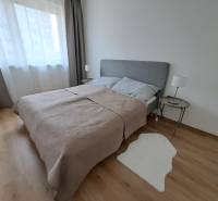 Hálószoba egy háromszobás lakásban, fa dekoros padlóval, szürke ágytámlával és szőnyeggel.