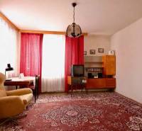 Nappali egy családi házban, piros szőnyeggel, fotellel és függönyökkel az ablakokon.