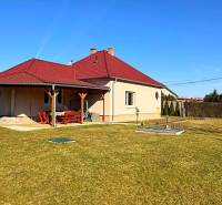 Családi ház Zemianska Olčán piros tetővel és fa előtetővel, füves kert.