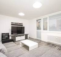 Nappali egy háromszobás lakásban kanapéval, televízióval és fa mintázatú padlóval.