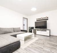 Nappali egy háromszobás lakásban kanapéval, TV-vel és fa mintázatú padlóval.