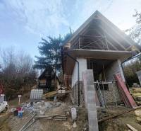 Nyúl-völgyi házikó építése Holčíkovce-ban, fákkal és építőanyaggal körülvéve.