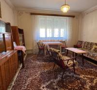 Nappali egy családi házban szőnyeggel, bútorokkal és csillárral, függönyök az ablakokon.