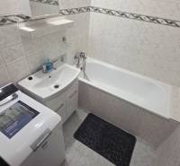 Fürdőszoba káddal és mosógéppel egy egyszobás lakásban, világos csempék és tárolóhely.