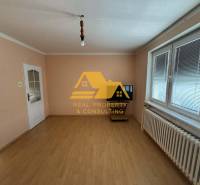 Egy családi ház szobája fehér falakkal, fa mintázatú padlóval és redőnyös ablakkal.