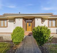 Családi ház Kútnikokban bokros ültetéssel, fehér homlokzattal és barna kiegészítőkkel.