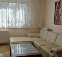 Világos nappali egy háromszobás lakásban kanapéval, absztrakt festménnyel és fa dekorációjú padlóval.