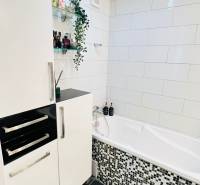 Fürdőszoba egy háromszobás lakásban fehér káddal, modern csempékkel és tárolóhellyel.