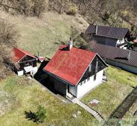 Ház Moštenicában ferde piros tetővel, környező zöld növényzet és lejtős terep.