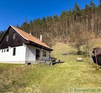 Házikó Moštenicában, füves területtel körülvéve, háttérben erdővel.