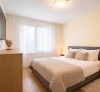 Hálószoba egy háromszobás lakásban szürke ággyal, fa mintázatú padlóval és falra szerelt TV-vel.