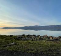 Nyugodt vízfelület, füves part és hegyek Vinné környékén. Lakóterületek.