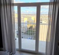 Garzonlakás kilátással az erkélyajtón keresztül, világos padló, függönyök.