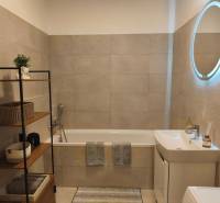 Kétszobás lakás fürdőszobája káddal, polcrendszerrel és világított tükörrel.