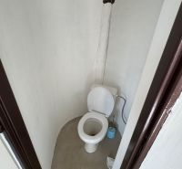 Egyszerű WC a házikóban, burkolat nélküli falakkal és műanyag WC-vel.