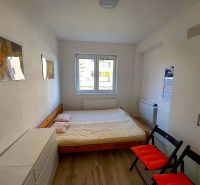 Garzonlakás ággyal, fehér szekrénnyel és fa mintázatú padlóval, két összecsukható székkel.