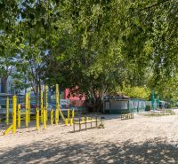 Park sárga edzőeszközökkel és fákkal a Budatínska utcánál, háttérben épületekkel.