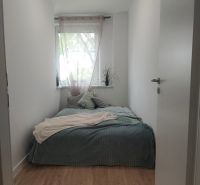 Keskeny hálószoba ággyal és ablakkal, fa dekoros padlóval, világos belső tér.