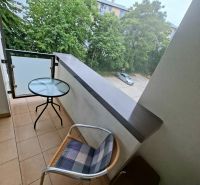 Garzonlakás erkélye kilátással a zöldre és parkolóra, szoba asztallal és székkel.
