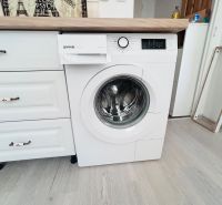 Mosógép az egyszobás lakás konyhájában, fa mintázatú padlóval és fa munkalappal.