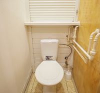 Kis fürdőszoba egy kétszobás lakásban WC-vel, fehér falakkal és oldalfalon lévő csövekkel.