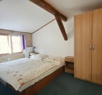 Hálószoba egy nyaraló apartmanban fa gerendákkal, szekrénnyel és ággyal az ablak mellett.