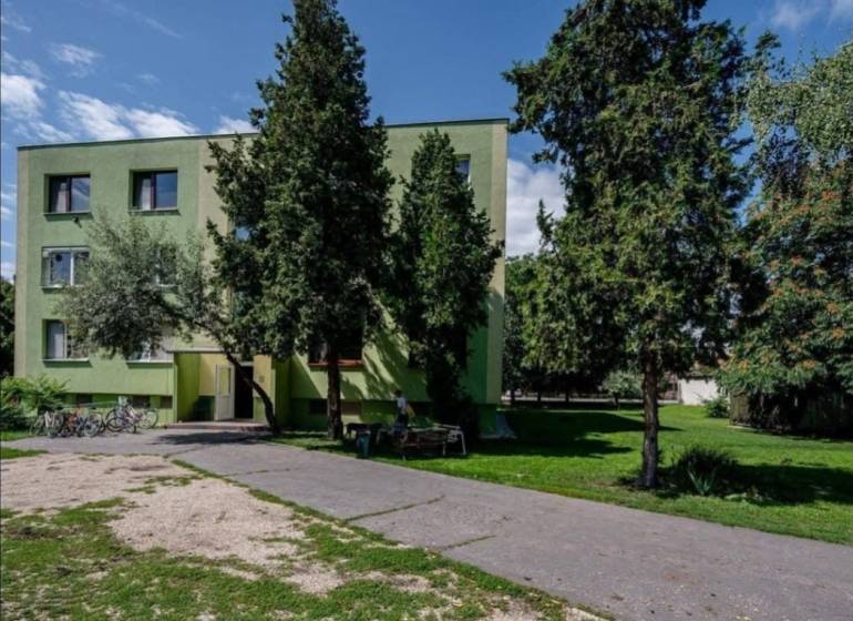 Egy fák által körülvett lakókonténer Csicsón, aszfaltút és kerékpárok mellett.