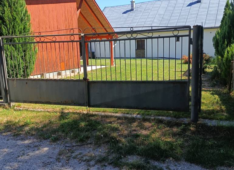 Kapú és faház Tužinában, gyep és fenyőfák veszik körül.