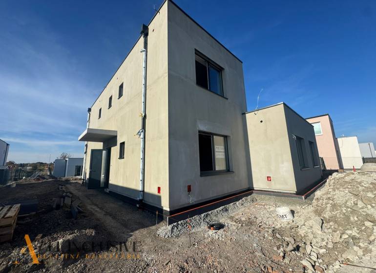 Hubicai családi ház építés alatt, minimalista külsővel és szomszédos épületekkel.