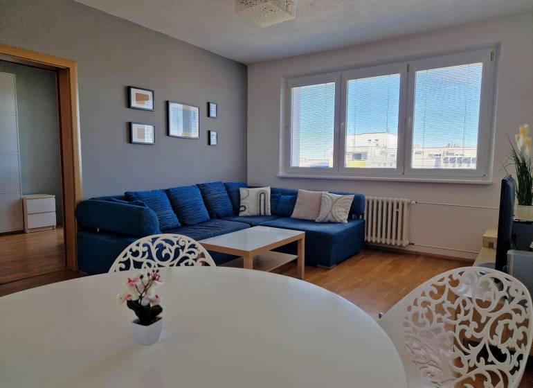 Nappali háromszobás lakásban kanapéval, fehér asztallal és fa dekorációjú padlóval.