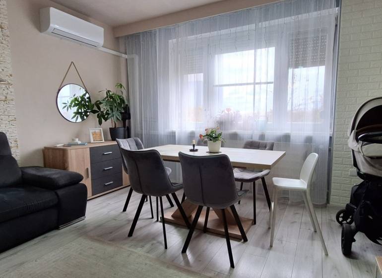 Nappali szürke kanapéval, fa dekorációs asztallal és finom függönyökkel egy háromszobás lakásban.