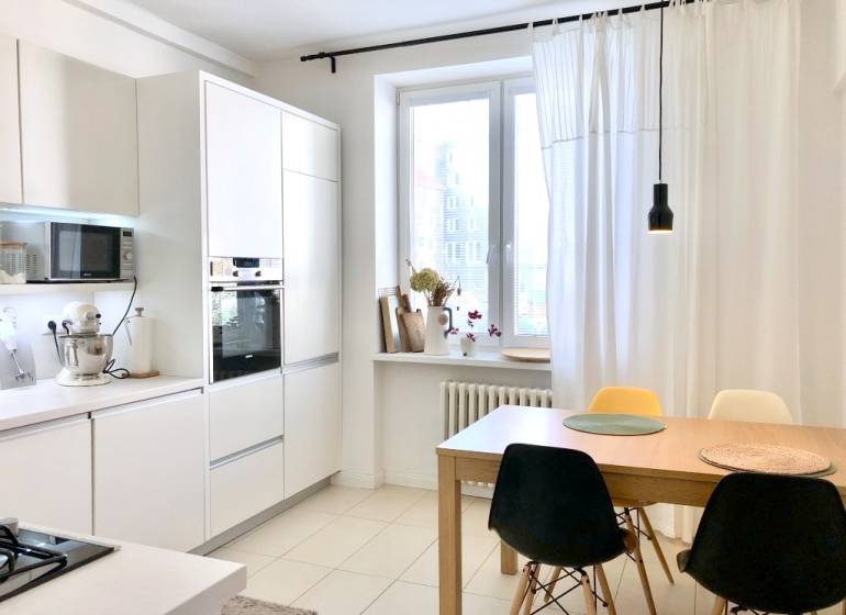 Konyha egy kétszobás lakásban fehér szekrényekkel és étkezőasztallal székekkel.