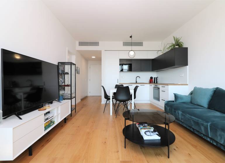 Nappali konyhával egy 2 szobás lakásban, fa dekorációjú padló, TV és ülőgarnitúra.