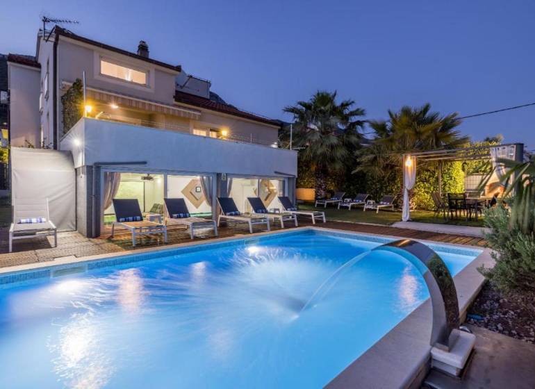 Villa Kaštelában megvilágított medencével, napozóágyakkal és pálmafákkal a kertben alkonyatkor.