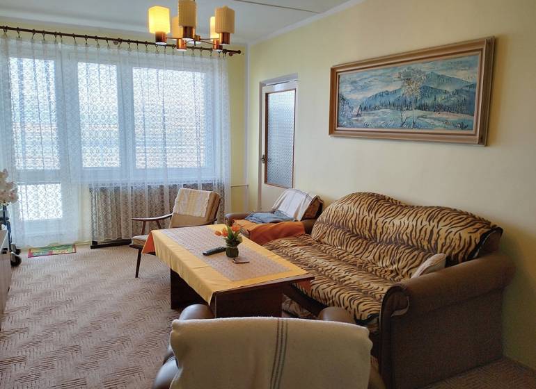 Nappali kanapéval, fotellel és asztallal egy kétszobás lakásban nagy ablakkal.