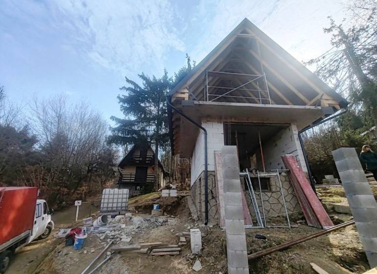 Nyúl-völgyi házikó építése Holčíkovce-ban, fákkal és építőanyaggal körülvéve.