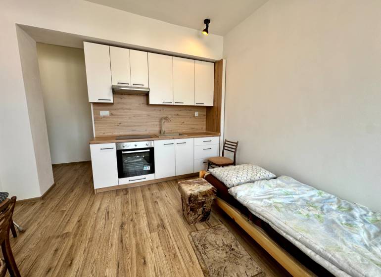 Garzonlakás konyhasarokkal és fa mintázatú padlóval, kiegészítve ággyal és székkel.