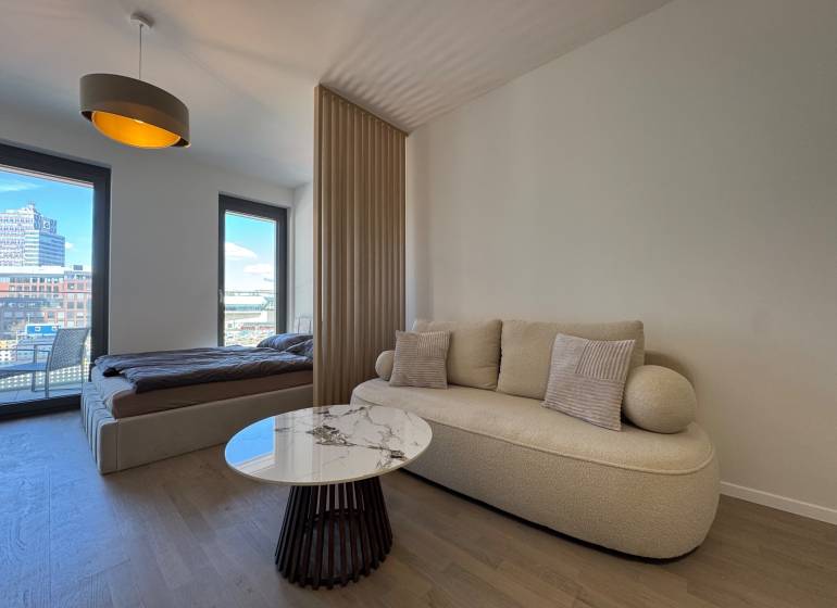 Nappali kanapéval, dohányzóasztallal és ággyal egy egyszobás lakásban, fa dekor padlóval.