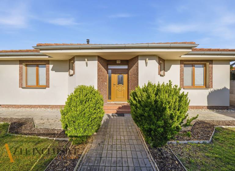 Családi ház Kútnikokban bokros ültetéssel, fehér homlokzattal és barna kiegészítőkkel.